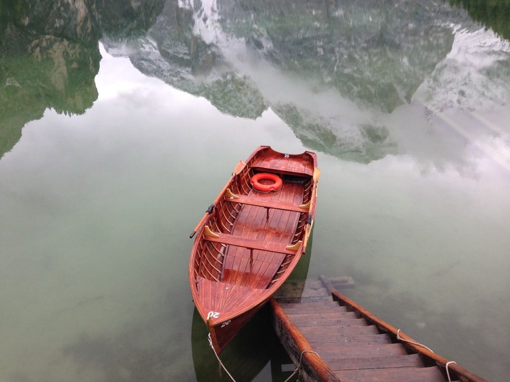 Dettaglio del lago di Braies con il riflesso della montagna di roccia la Croda del Becco, in primo piano la scaletta che scende dal pontile per raggiungere le barche e una barchetta ormeggiata con un salvagente rosso appoggiato sul fondo.