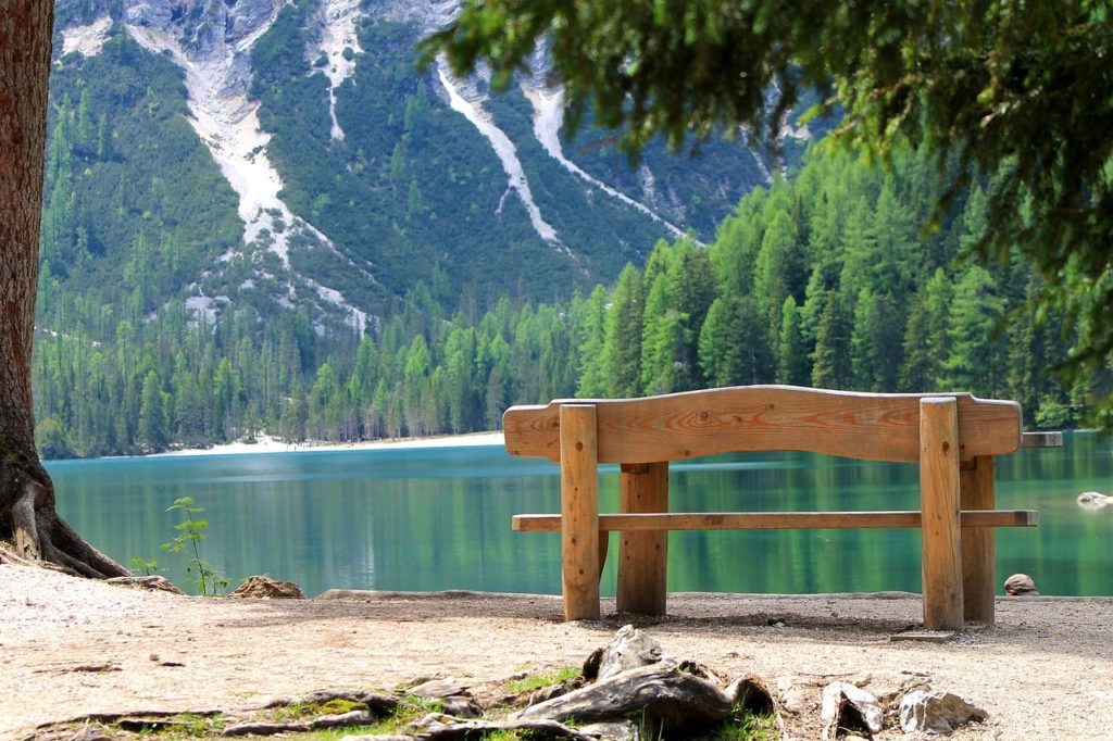 Scorcio del lago di Braies con una panchina in primo piano, l'acqua verde che riflette pini e abeti e sullo sfondo i canaloni ancora innevati della montagna rocciosa